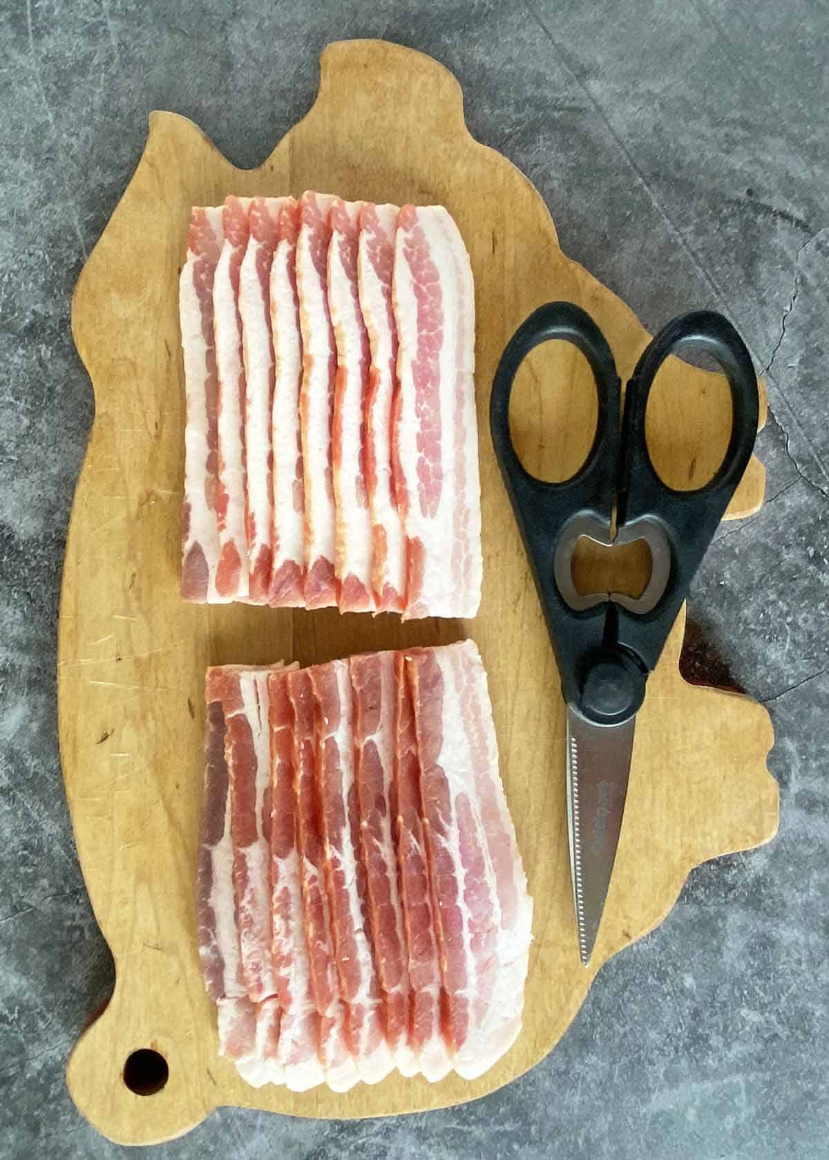 A cutting board with a pound of bacon and a pair of kitchen shears.