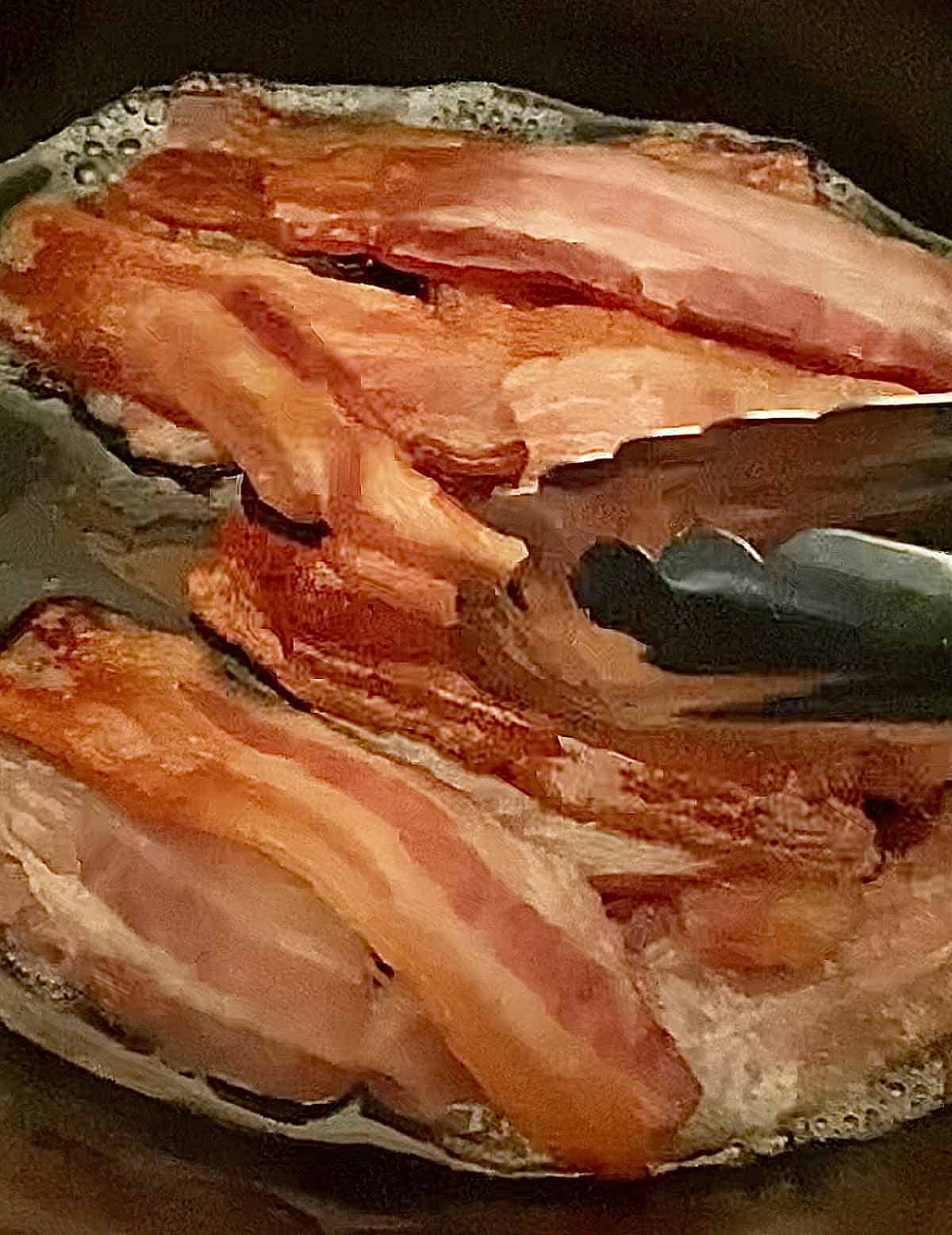 A pair of tongs rearranging partially cooked bacon slices in a slow cooker.