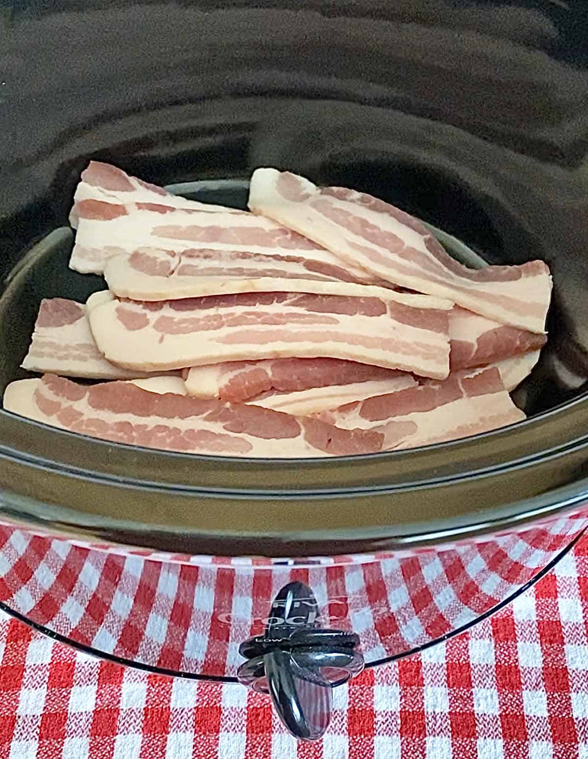 8 halved slices of bacon in the bottom of a black slow cooker appliance.