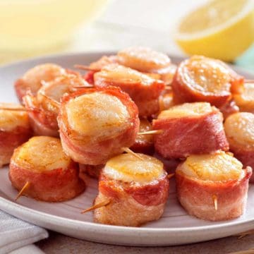 A dozen bacon wrapped scallops on a white serving dish with a cut lemon in the background.