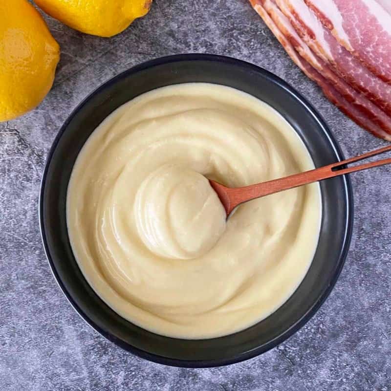 Black serving bowl filled with bacon aioli with lemons and bacon in the background.