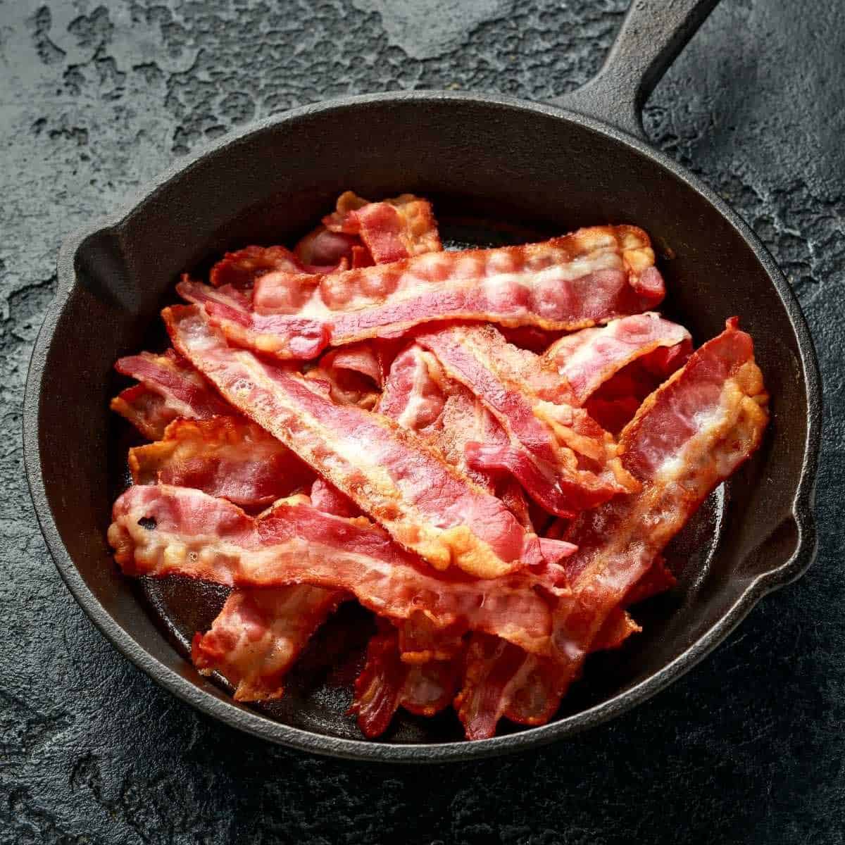 A pound of cooked bacon strips in a black skillet on a mottled black background.