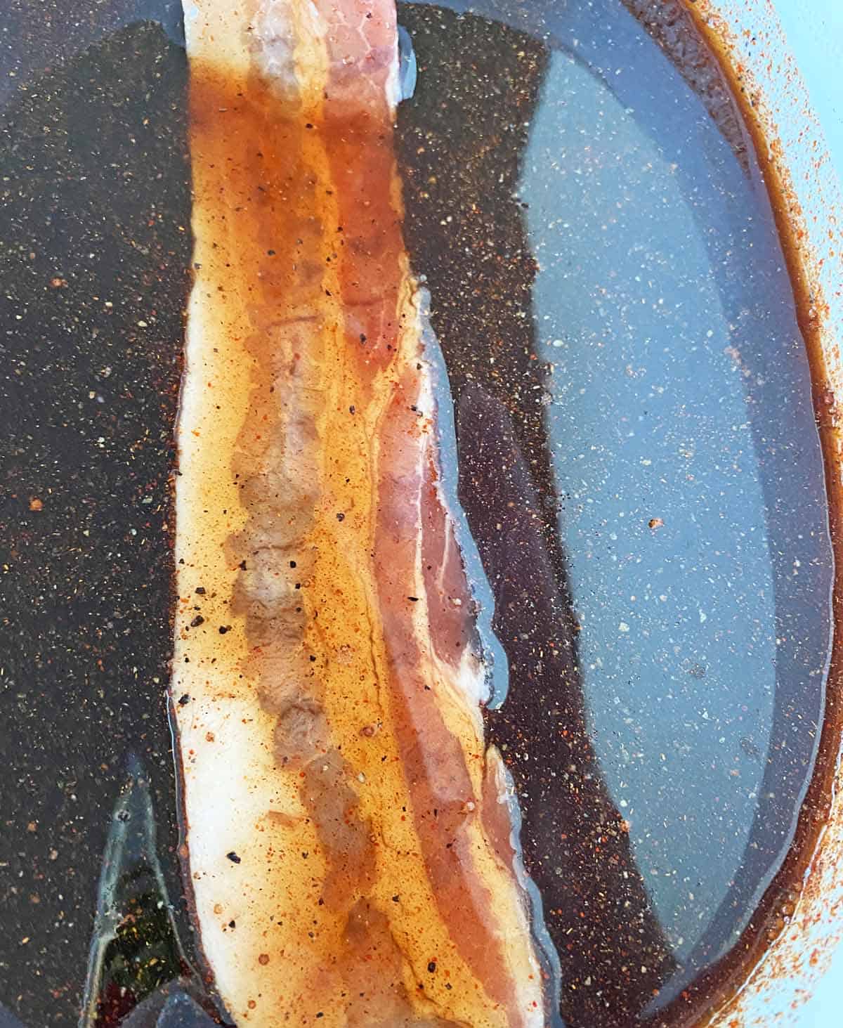 One slice of thick sliced bacon being dipped in million dollar bacon glaze in a white dish.