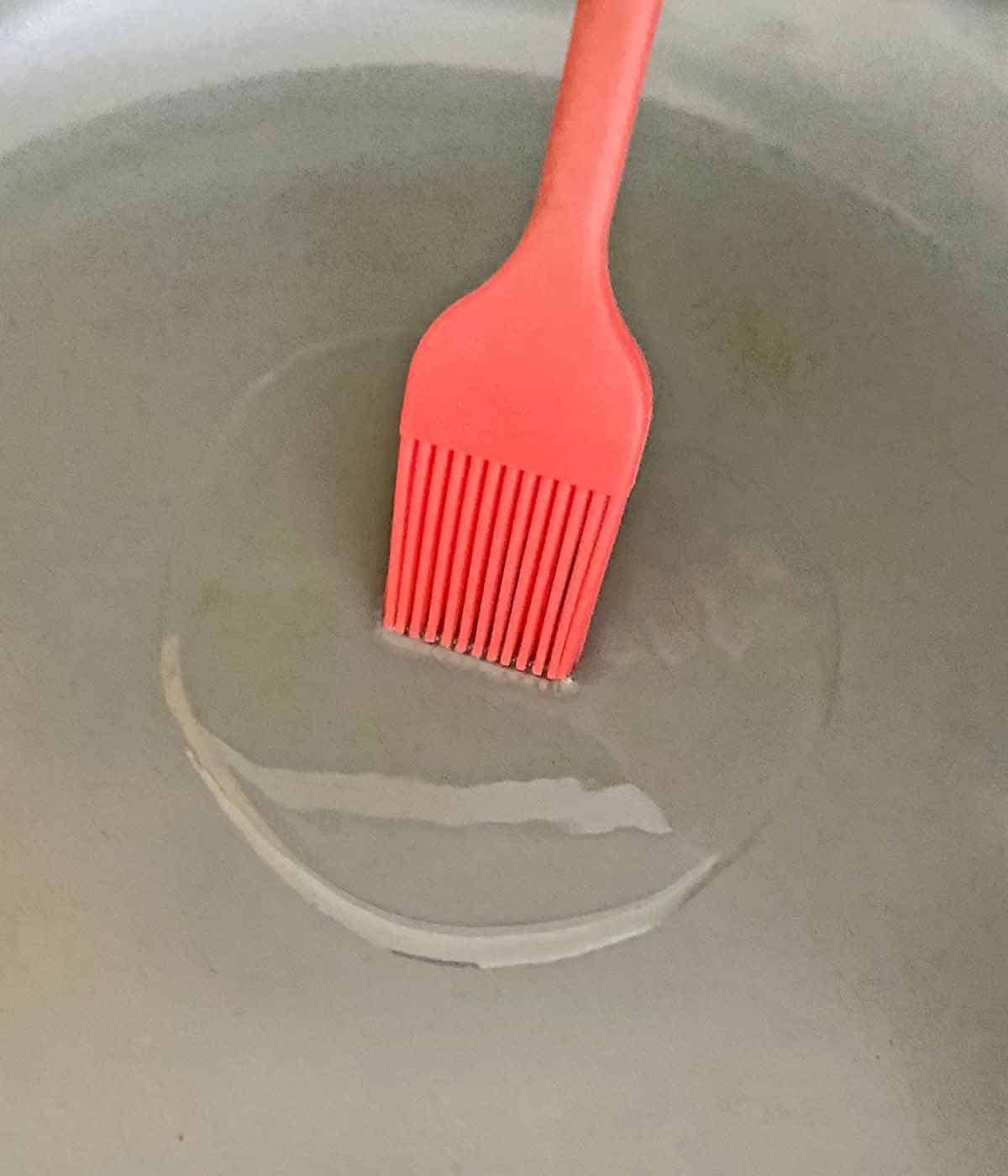 A red silicone brush spreading oil in a frying pan.