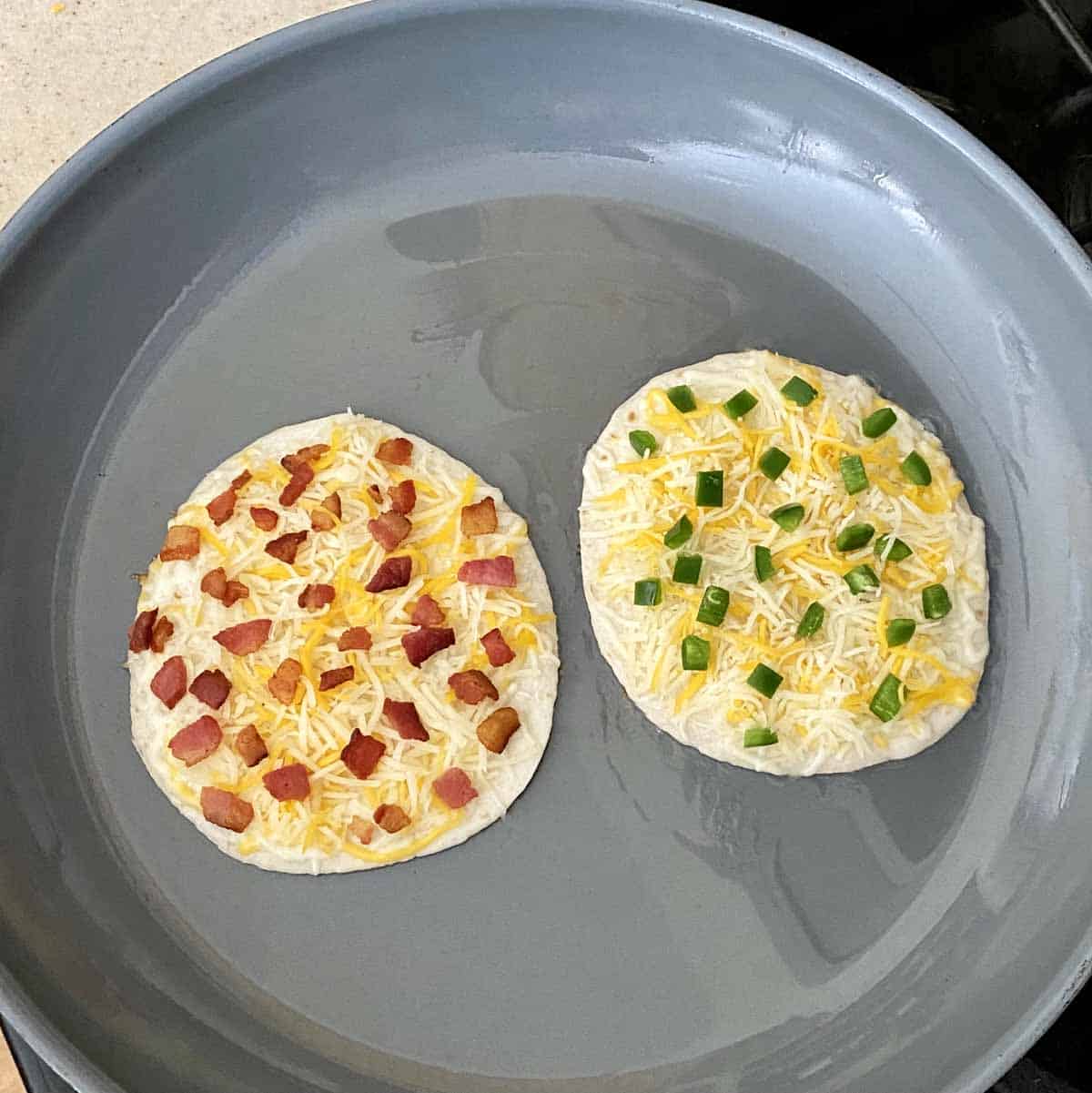 Two mini flour tortillas topped with cheese, bacon and chopped jalapeños just starting to cook in a hot skillet.