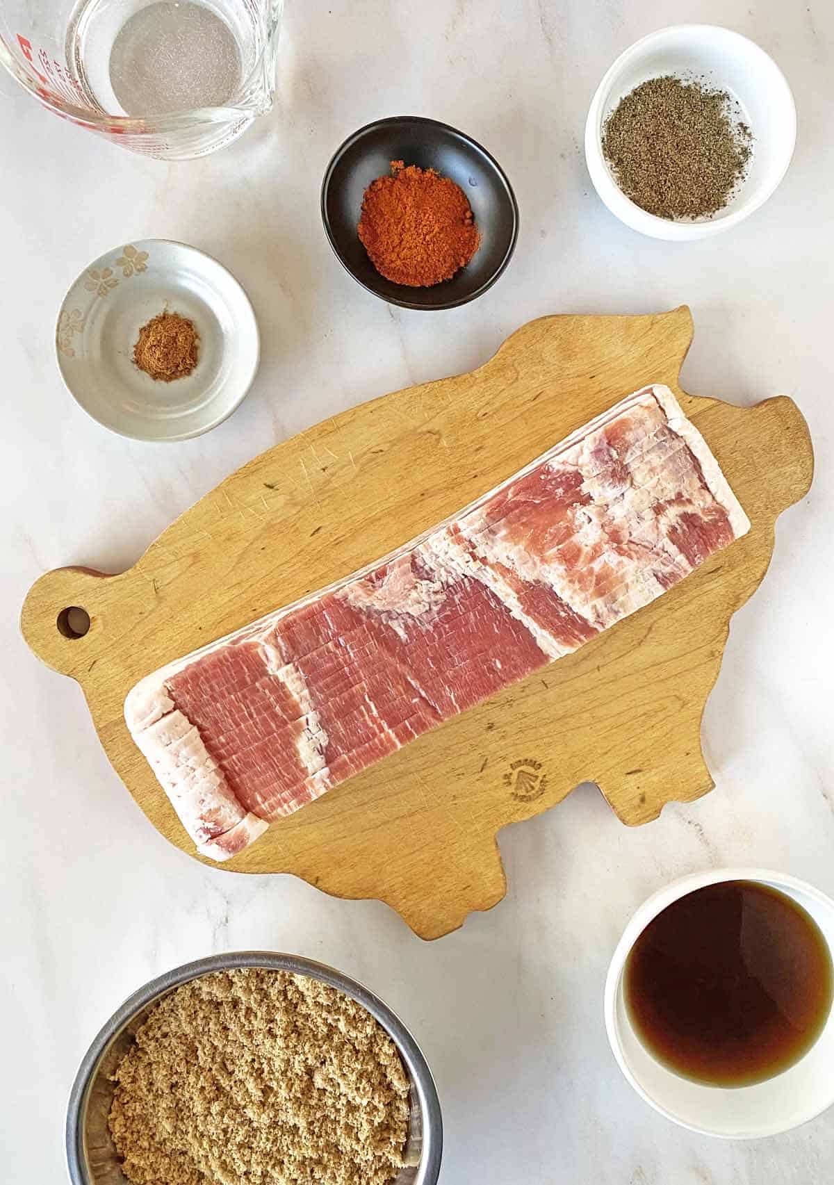 Spices, a measuring cup of water, a slab of bacon on a cutting board, brown sugar and maple syrup for Million Dollar Bacon.