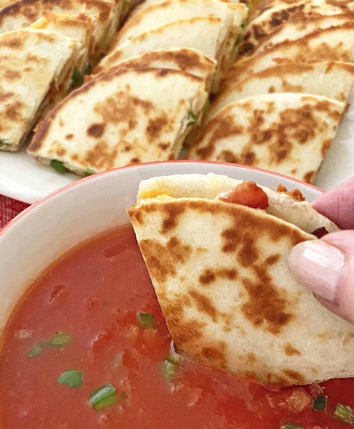 Dipping a quartered mini quesadilla with bacon and cheese in a bowl of salsa, with more quesadillas in the background on a platter.