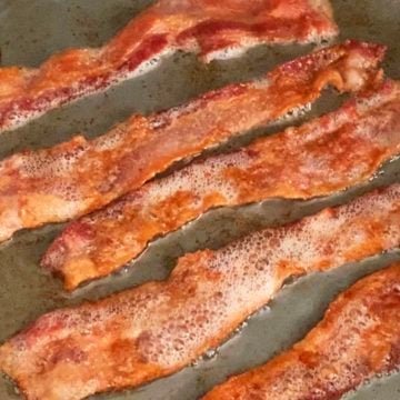 five strips of bacon cooking with fat in a skillet.