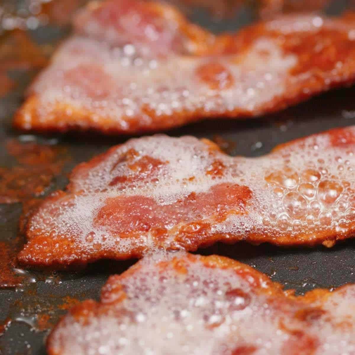 Close up of 3 strips of bacon cooking in a skillet and rendering bacon grease.