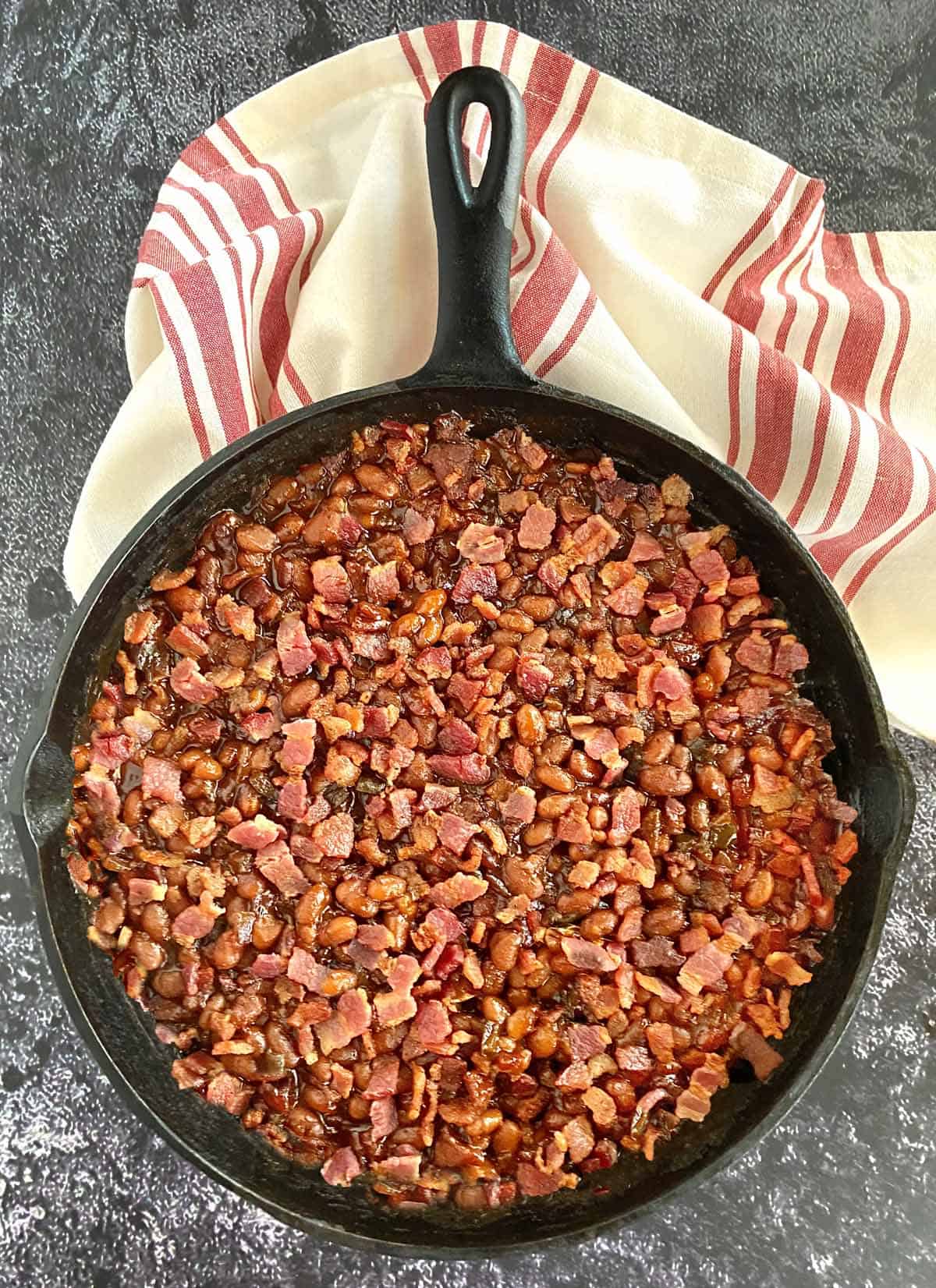 A cast iron skillet filled with smoked baked beans with bacon and brown sugar, with a cloth napkin near the handle.