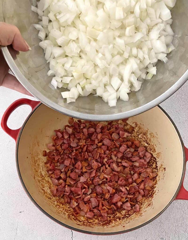 Adding chopped onion to the cooked bacon in a large pot with red handles.