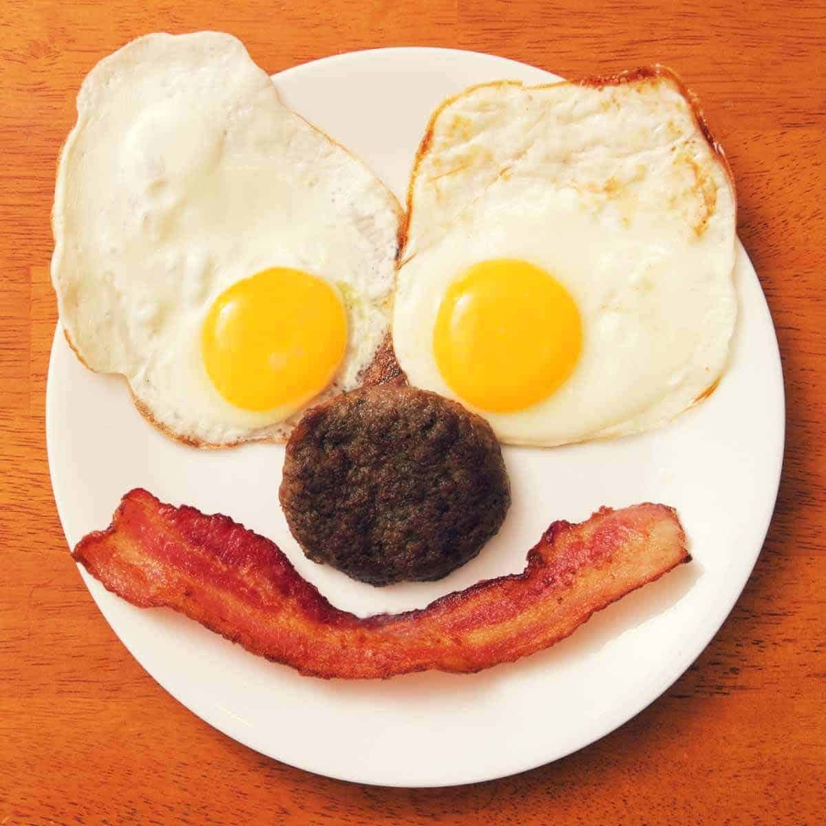 Two fried eggs, a round piece of fried sausage, and a strip of fried bacon arranged on a plate like a smiling face.