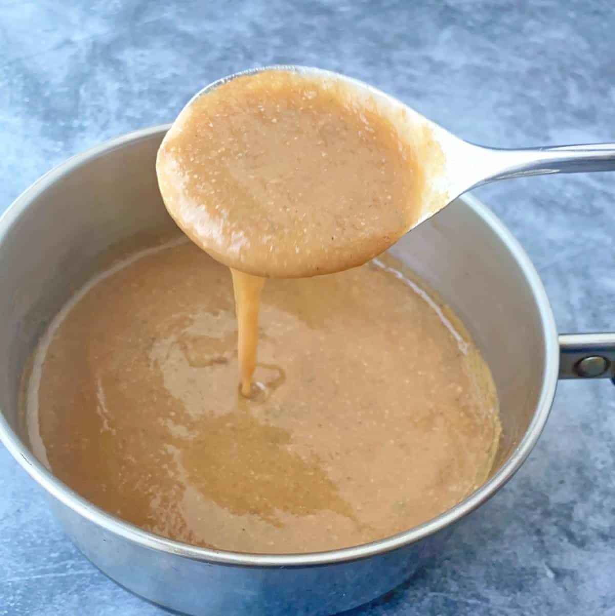 A silver spoon dipped in a saucepan of bacon gravy, with gravy dripping off the spoon.