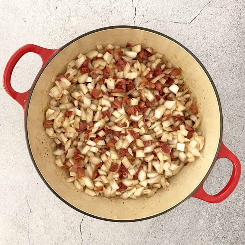 Onions and bacon cooking over low heat in a pot with red handles.