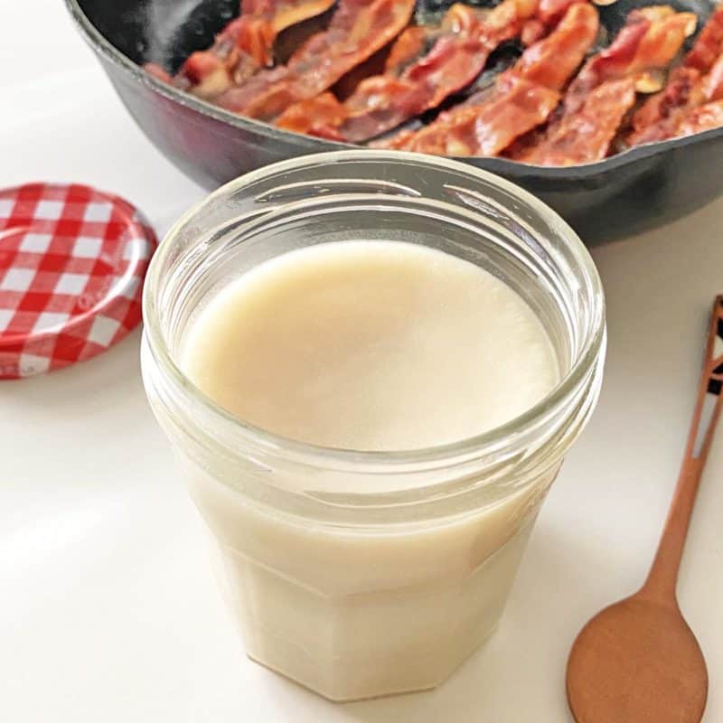 An open glass jar with chilled rendered bacon fat in front of a frying pan with six strips of golden brown cooked bacon.
