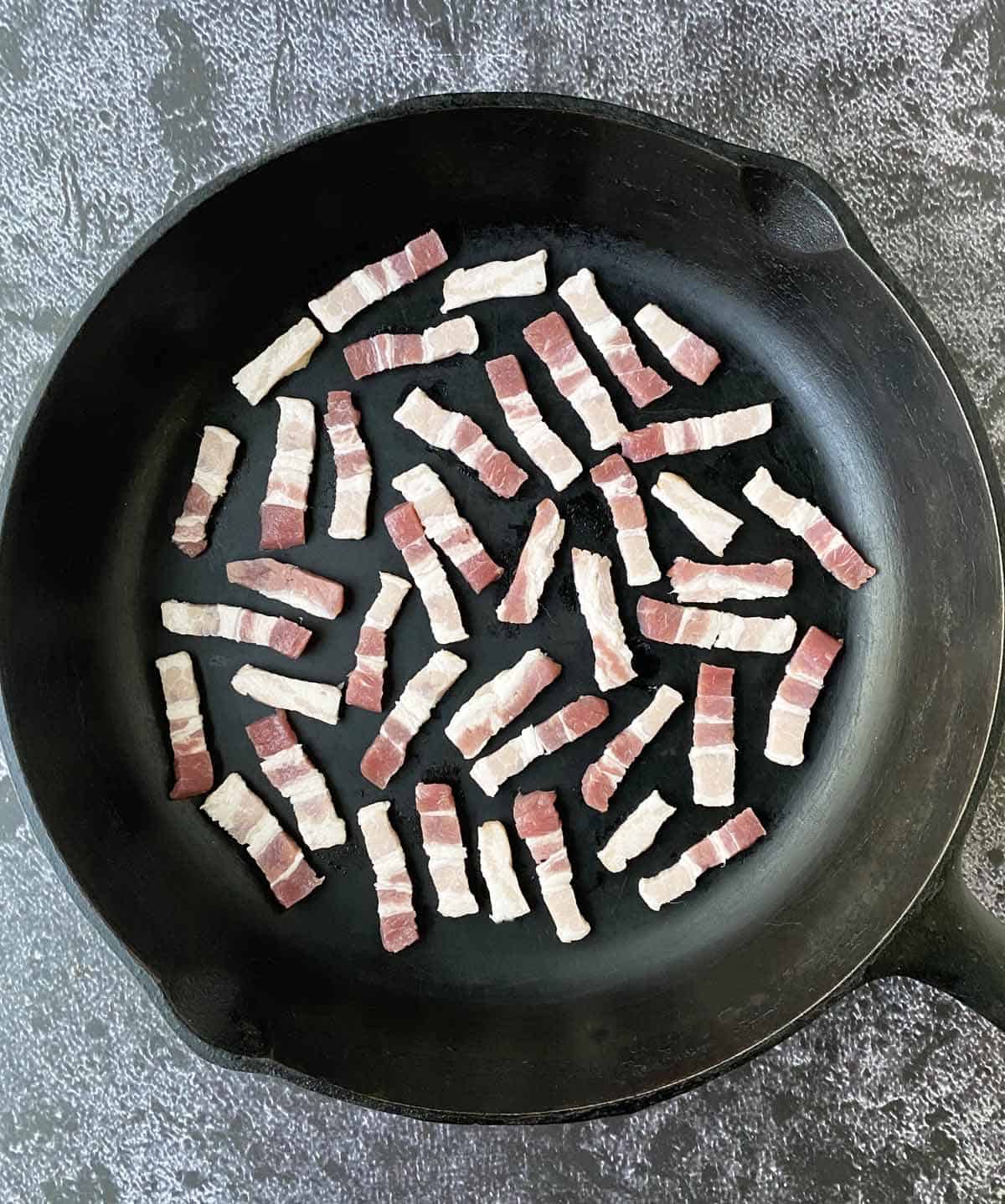 Several dozen raw bacon lardons in a cold cast iron skillet.