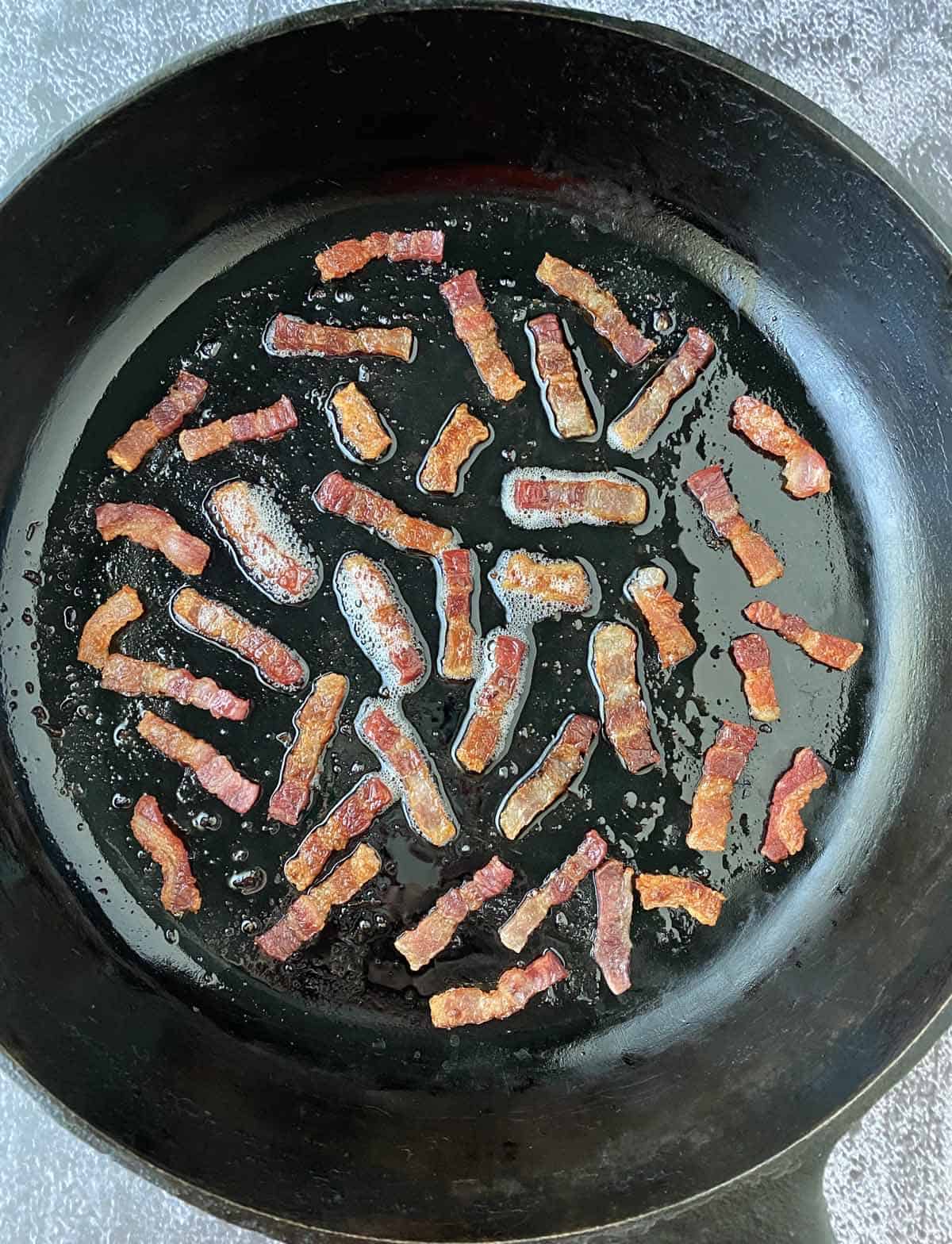 Bacon lardons cooking in a black cast iron skillet with rendered bacon fat.