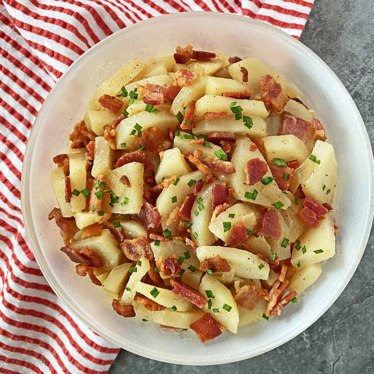 Warm potato salad with bacon garnished with chives in a white bowl on a read and white striped napkin.