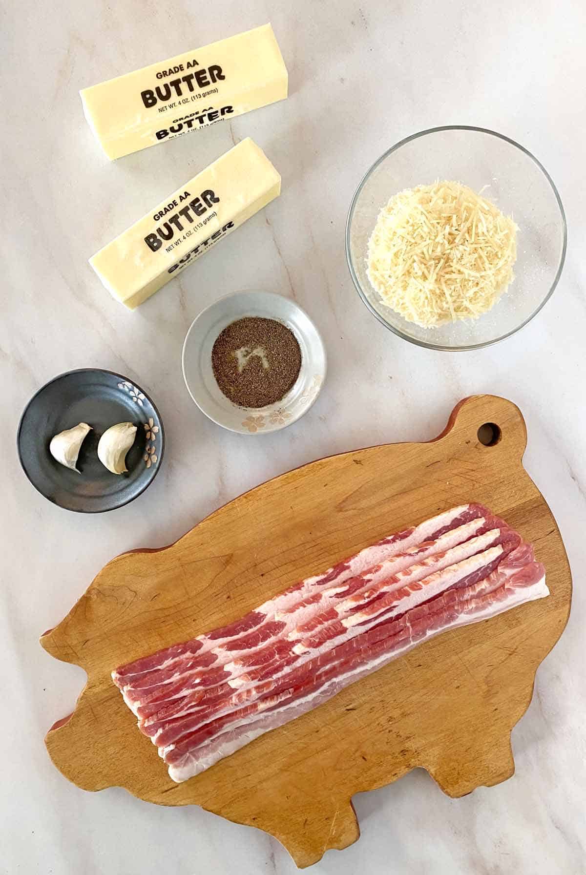 Two sticks of butter, a bowl of shredded Parmesan cheese, ground black pepper, two garlic cloves, six bacon slices and a pig shaped cutting board.