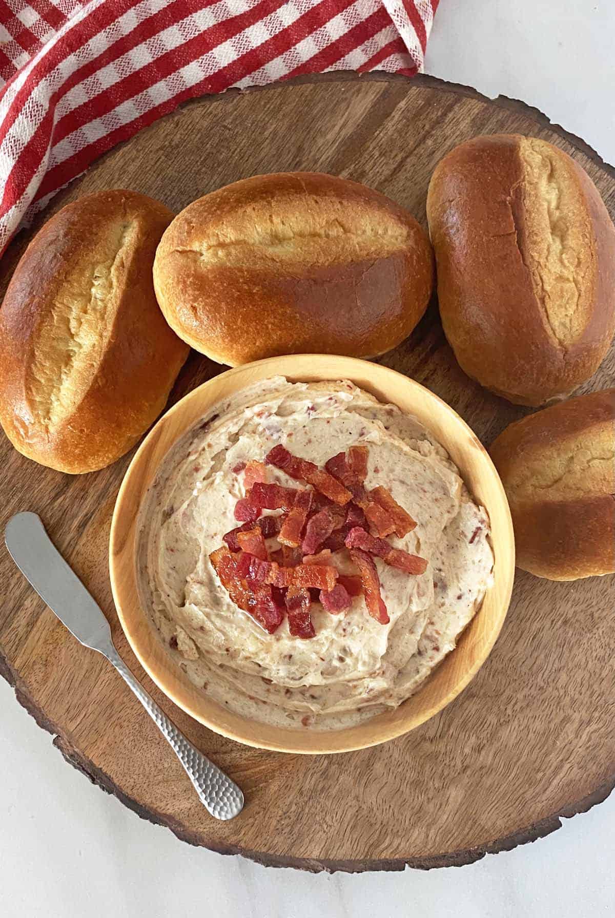 A bowl of bacon butter garnished with bacon lardons, four rolls, and a spreader knife on a wood background.