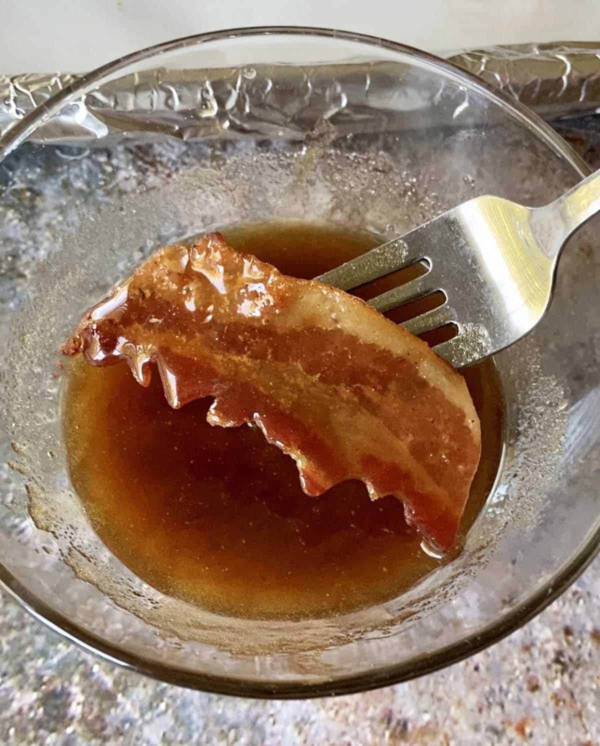 A fork with a bat shaped piece of bacon dipping it in the brown sugar maple syrup glaze in a small glass bowl.