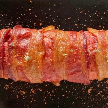 Smoked pork tenderloin wrapped in bacon, sprinkled with rub and on a black serving plate.