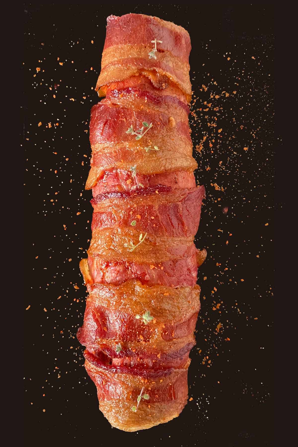 Fully cooked smoked pork tenderloin wrapped in bacon, sprinkled with rub and served on a black platter.