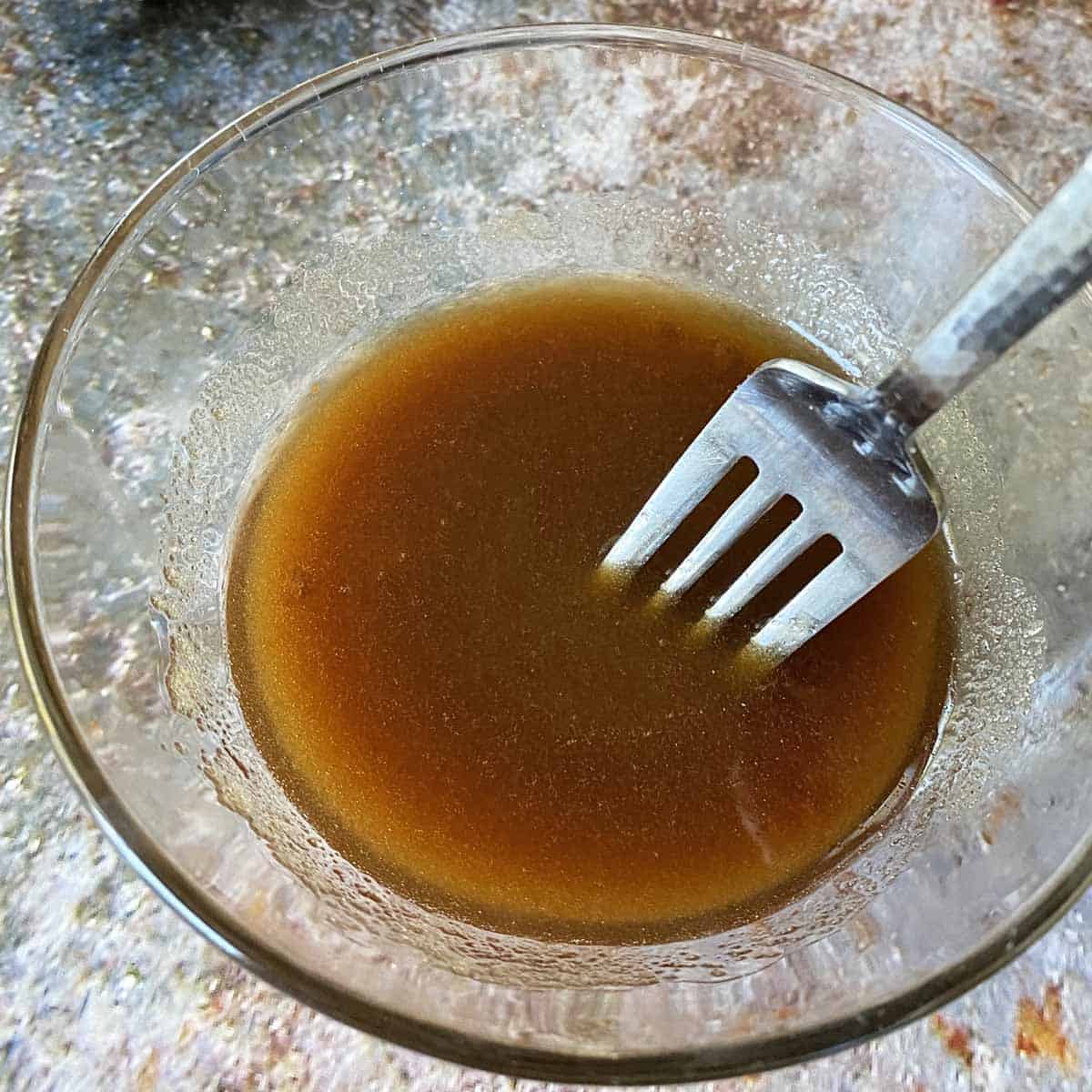 A fork stirring maple syrup and brown sugar together to make glaze, in a glass bowl.