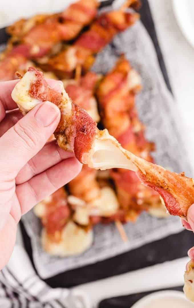 A hand holding a bacon wrapped mozzarella stick with more in the background.
