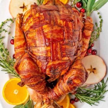 Bacon wrapped turkey on a platter surrounded by fresh herbs, oranges and apple slices.