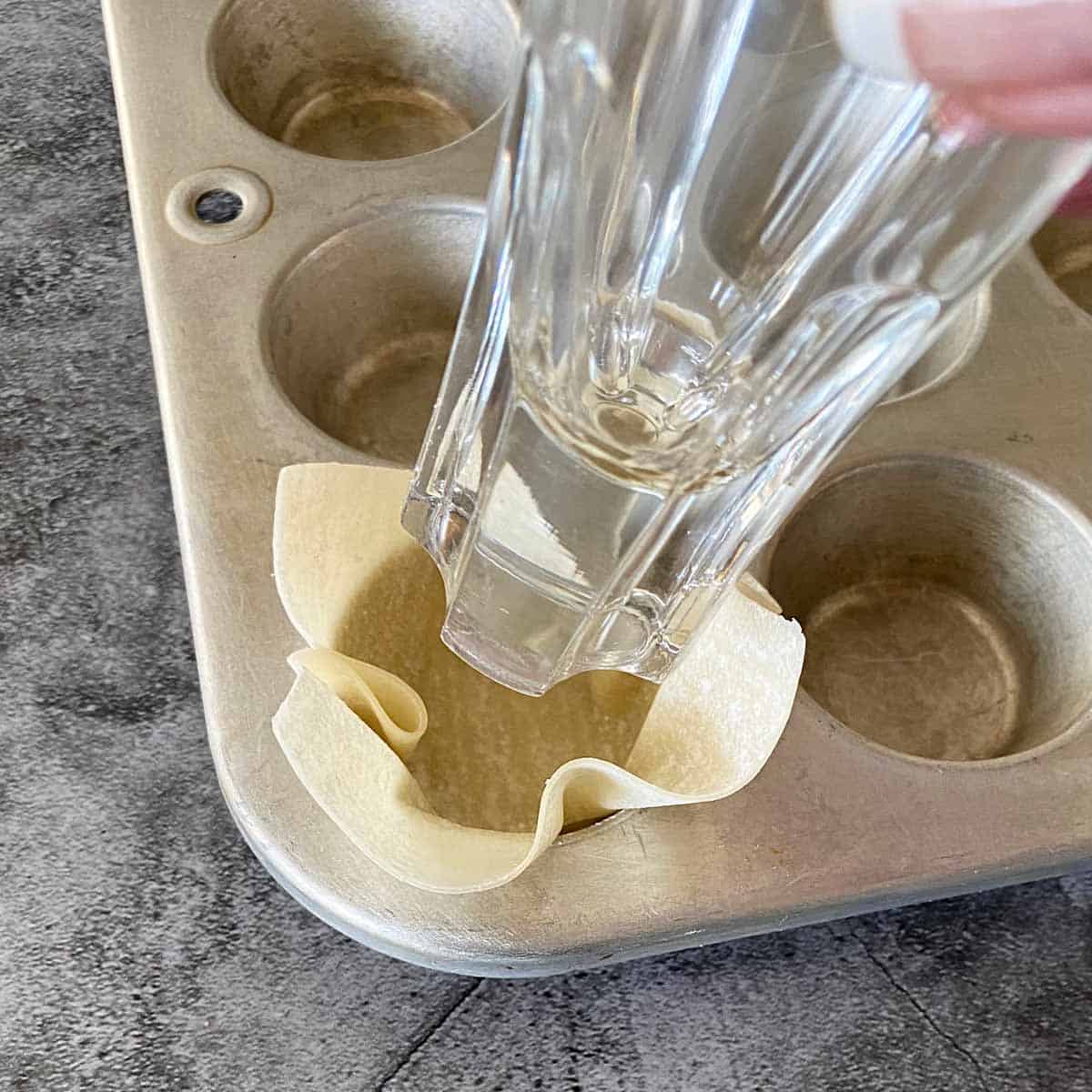 Using a shot glass to press a wonton wrapper in a mini muffin tin to make a cup shape.