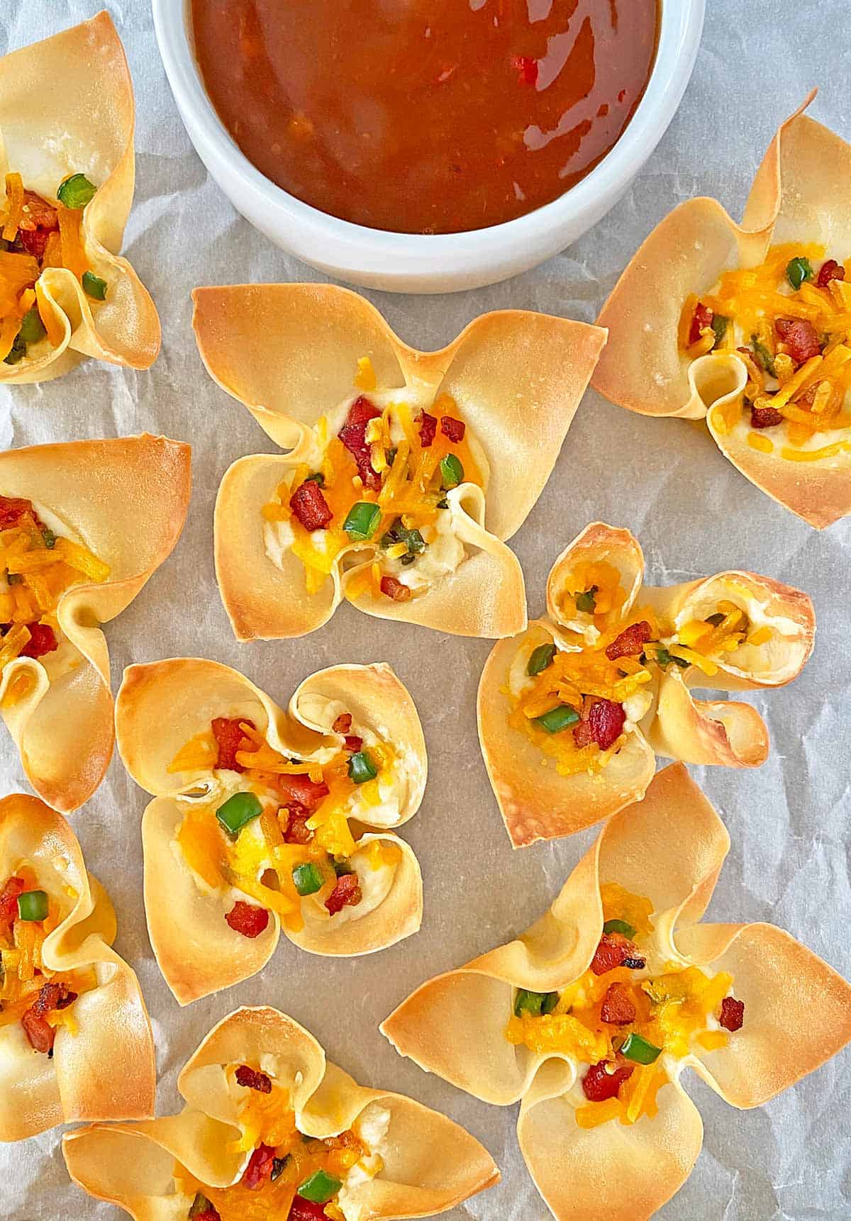 Crispy golden brown wonton appetizers with jalapeno popper filling on parchment paper with a bowl of sweet chili sauce for dipping.