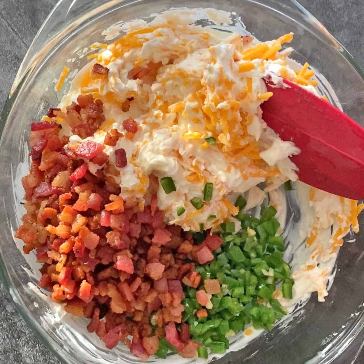 Stirring cooked bacon bits, jalapeno, cheddar cheese, sour cream and cream cheese with a red spatula in a glass bowl.