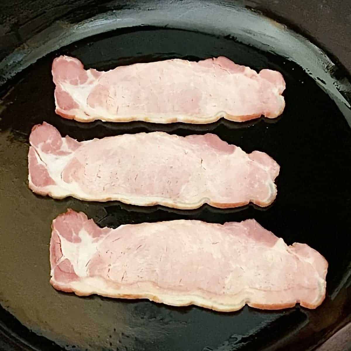 Three strips of back bacon in a black cast iron skillet.