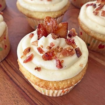 A maple bacon cupcake with vanilla bean buttercream frosting and candied bacon garnish on a wood board, with more cupcakes in the background.