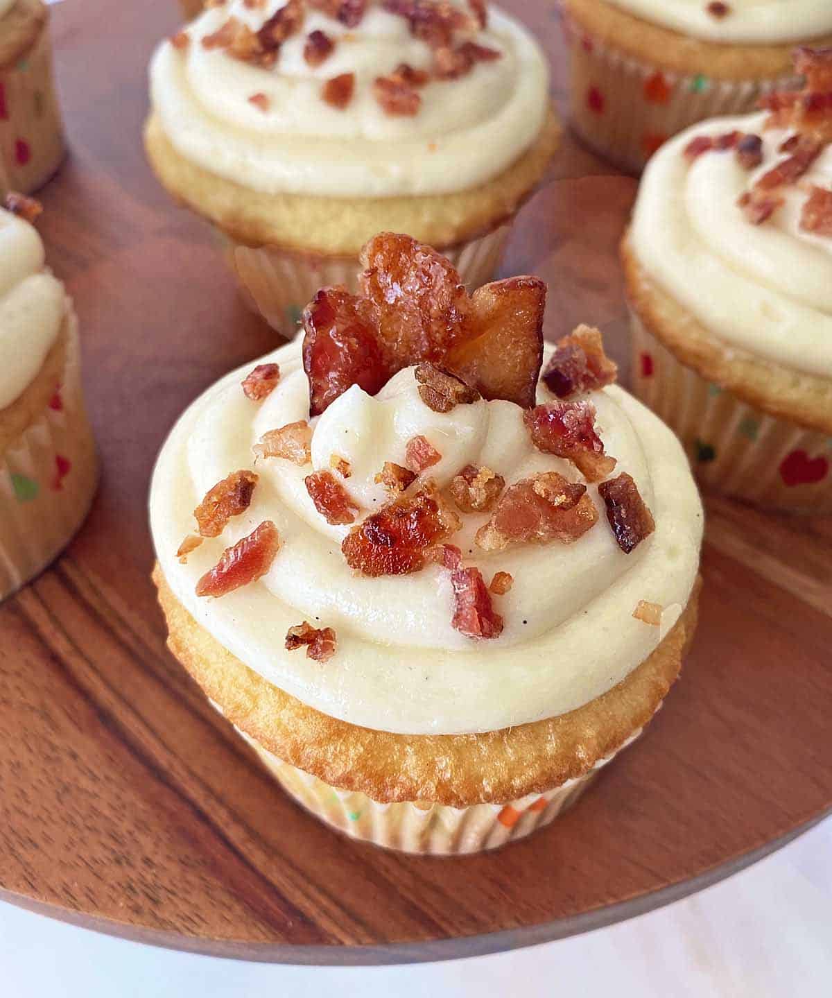 A finished maple bacon cupcake with vanilla buttercream frosting and candied bacon garnish on a wooden cake stand, with more cupcakes in the background.