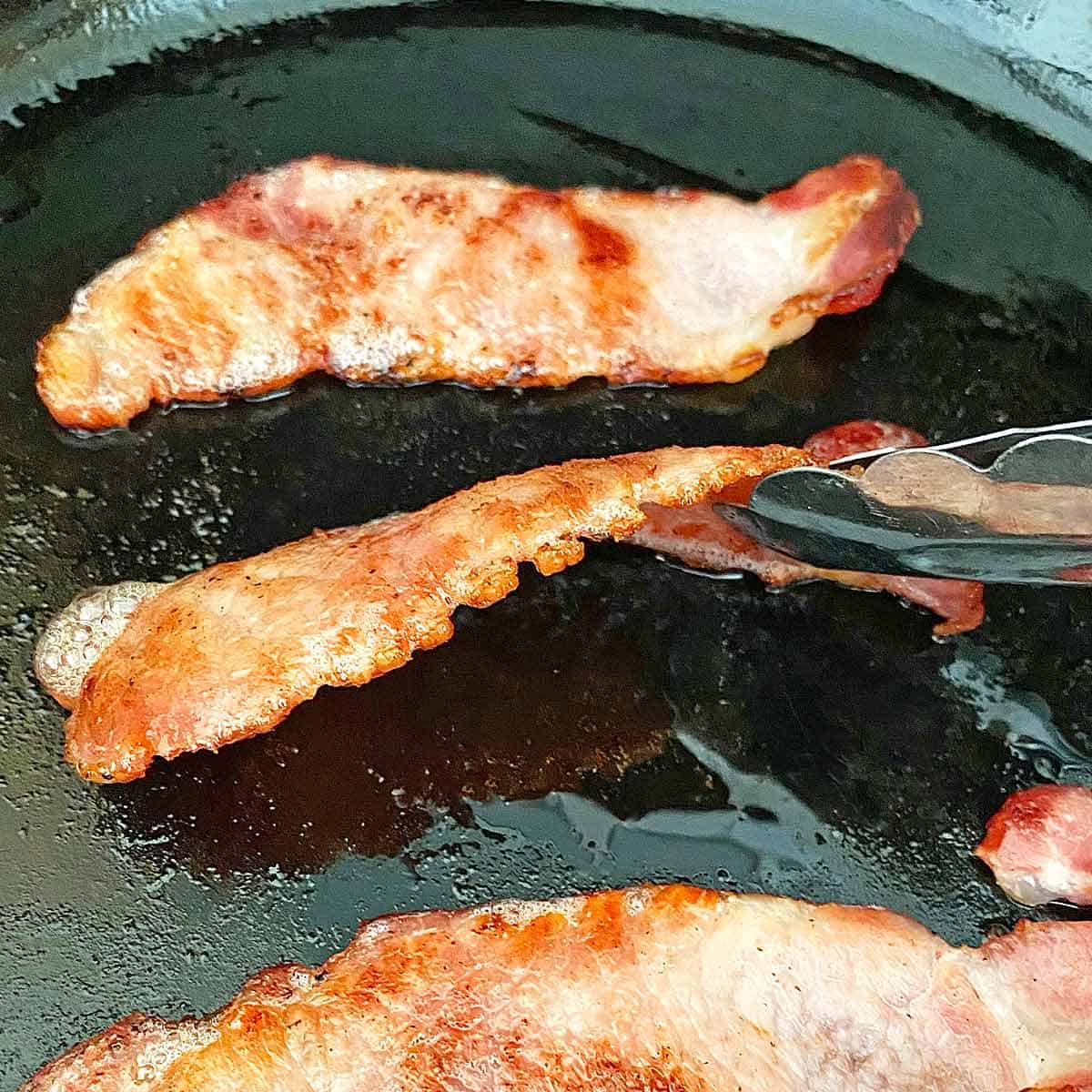 3 strips of back bacon in a skillet that have been notched on the edge to prevent curling.