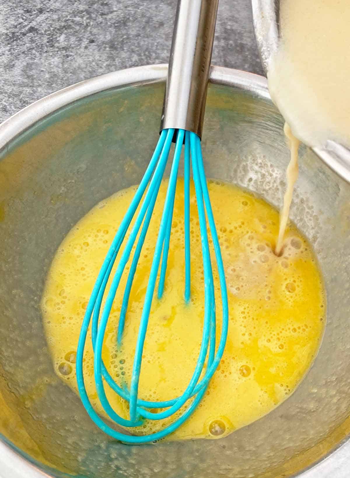Blending the milk mixture into the beaten eggs with a blue whisk.