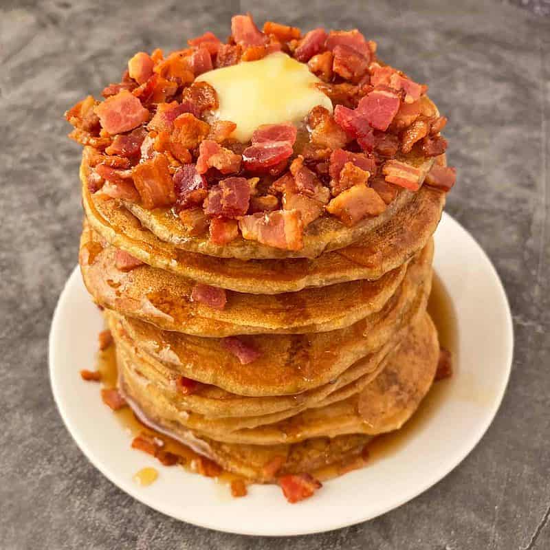 Bacon pancakes with maple syrup stacked on a white plate and topped with bacon bits and butter.