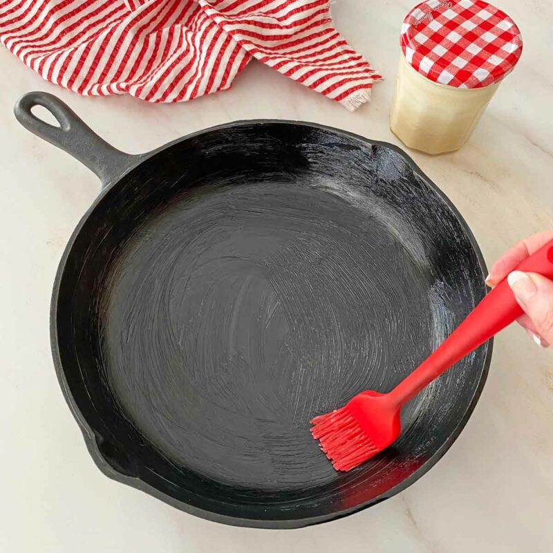 Brushing bacon grease on a cast iron frying pan with a red pastry brush.