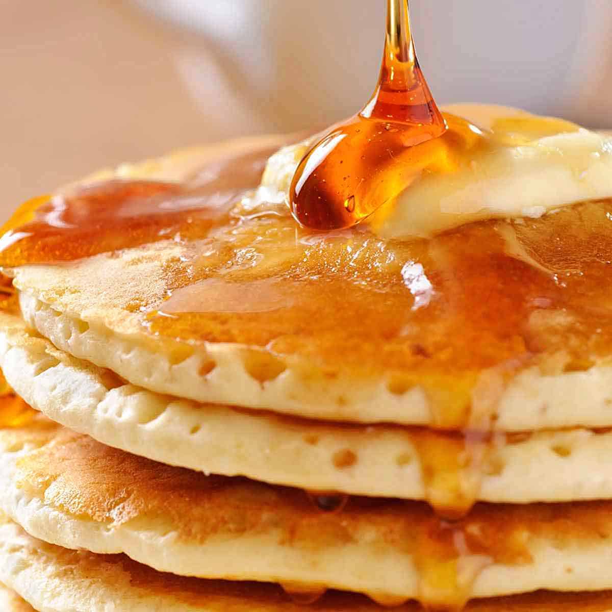 Four large pancakes with butter on top, being drizzled with maple syrup.