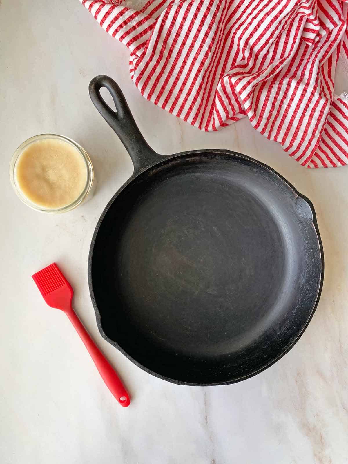 A cast iron pan, a brush, a jar of bacon fat and a red and white towel.