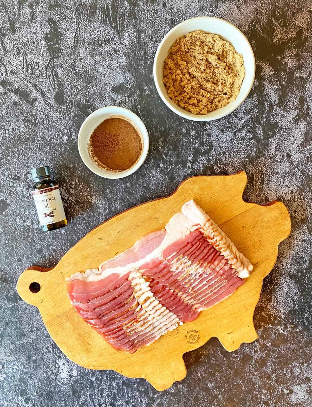 Brown sugar, ground cinnamon, cinnamon oil and sliced bacon on a pig shaped cutting board.