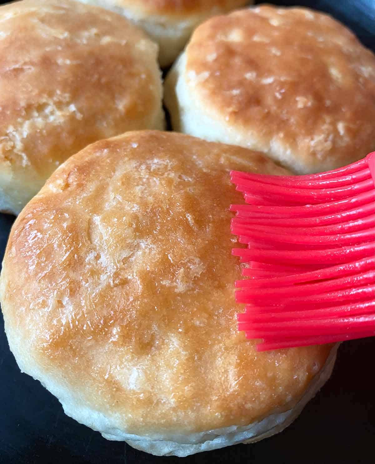 Brushing warm biscuits with melted butter using a red pastry brush.