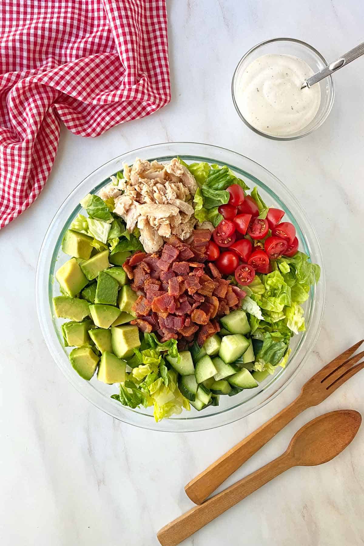 A large bowl of chicken bacon ranch salad, wooden salad tongs, a bowl of Balsamic ranch salad dressing and a red and white napkin.