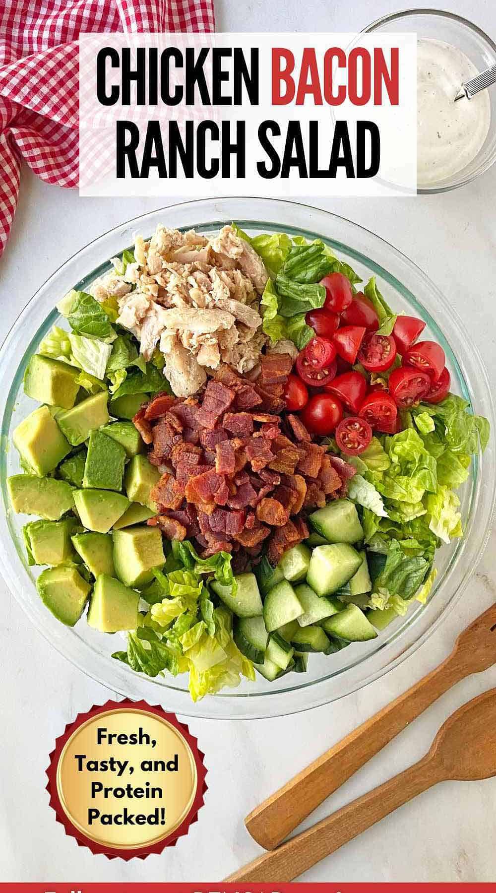 A glass bowl filled with fresh salad topped with bacon, tomatoes, avocado, cucumbers and chicken.