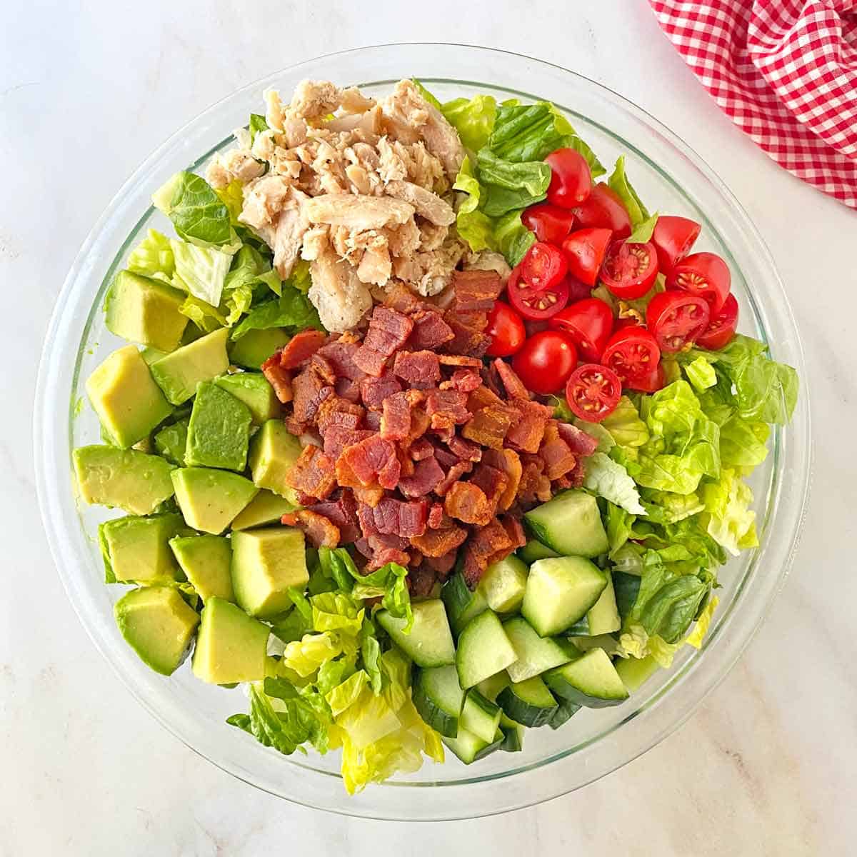 Finished salad with bacon, tomatoes, cucumber, chicken, avocado and ranch dressing in a glass bowl.