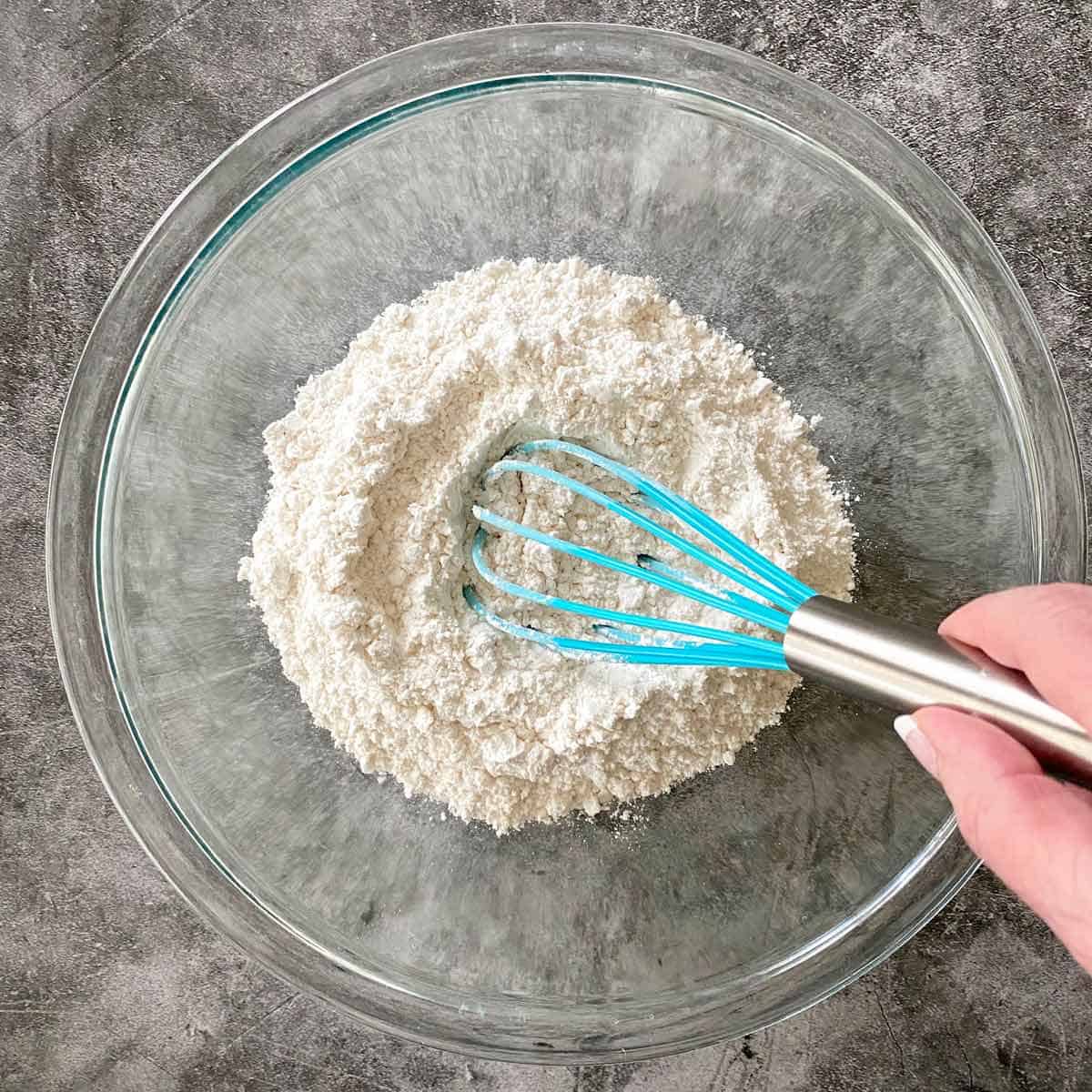 whisking dry ingredients together in a glass bowl with a blue whisk.