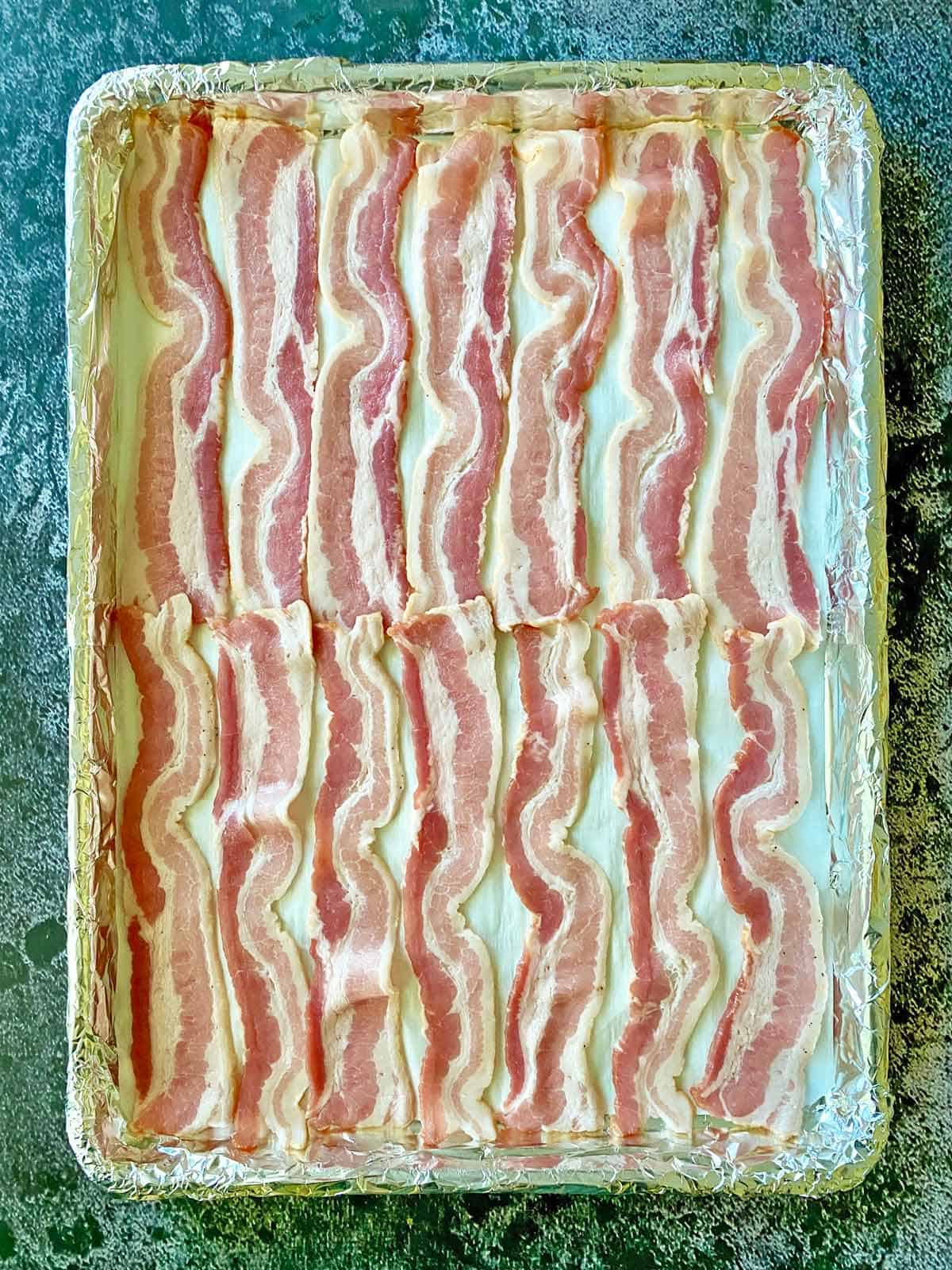 Uncooked bacon strips on a foil lined baking sheet.