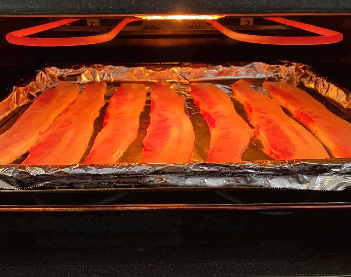 A foil lined baking pan with bacon slices being placed under an oven broiler.